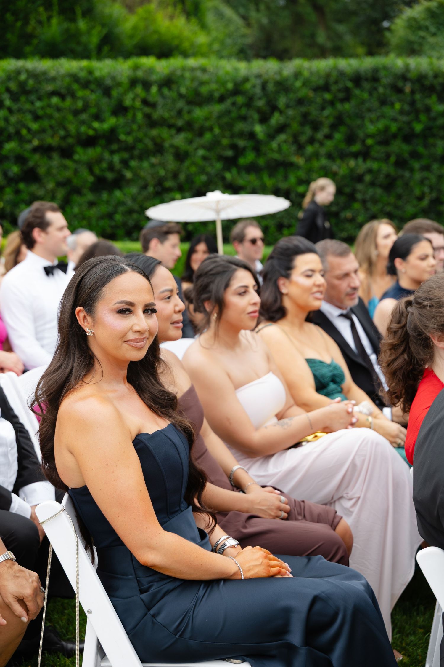 Elegant wedding guests seated outdoors in white chairs, woman in navy strapless gown smiling, lush green hedge backdrop.