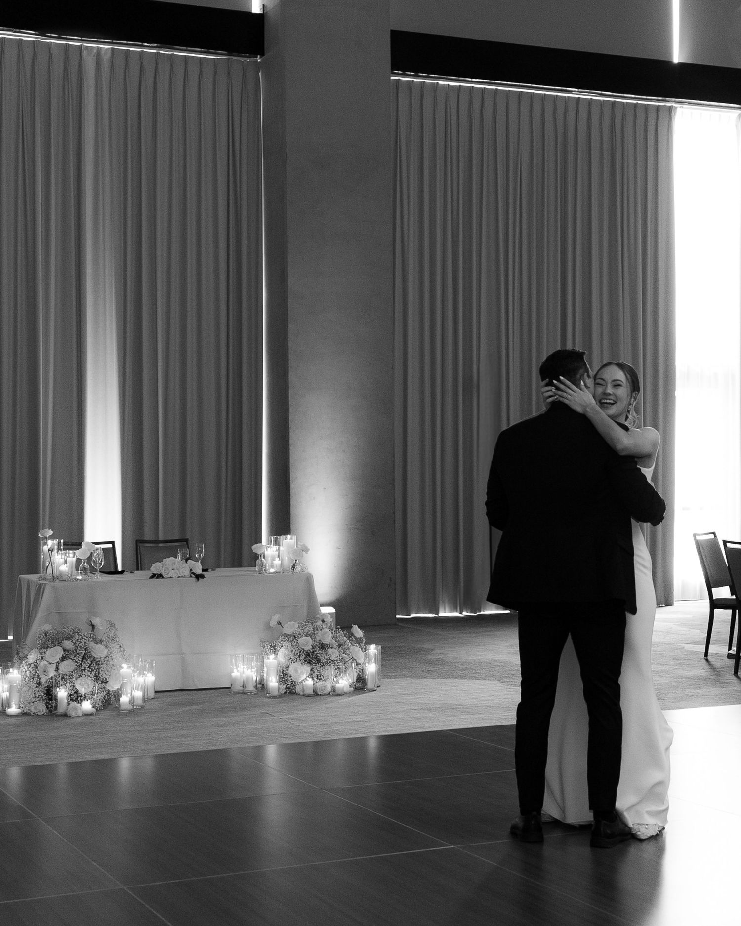 A black and white panoramic photo of couples dancing in a dimly lit ballroom with elegant curtains and decor.