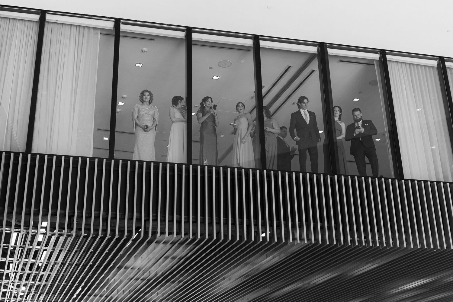A black and white architectural photograph showing a diagonal line of people standing behind glass windows.