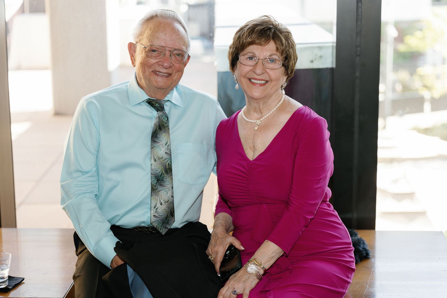 Two seniors in bright colored clothing sit together and share a laugh on an outdoor porch.