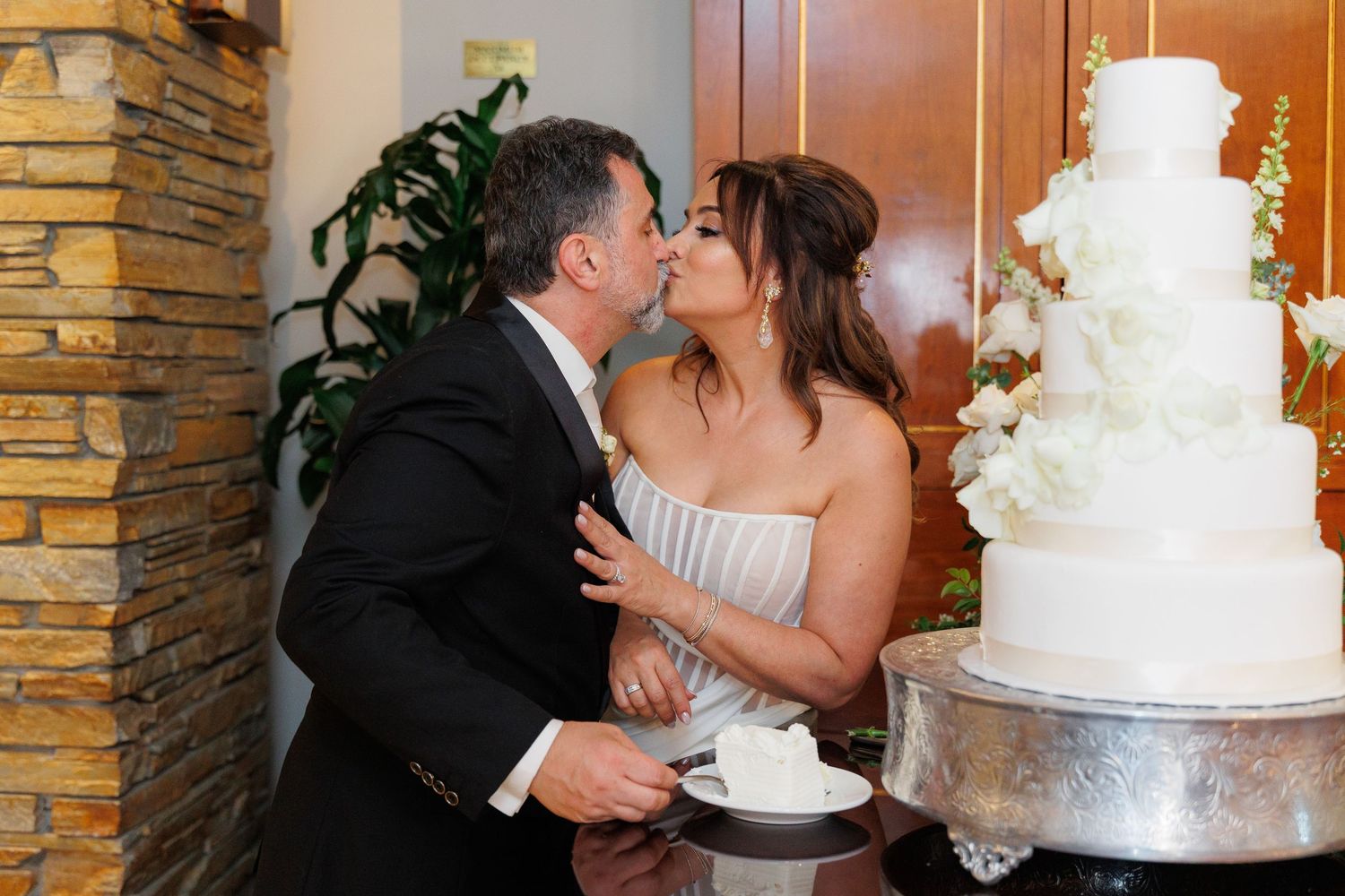 A couple shares a kiss next to an elegant white tiered wedding cake at their reception.