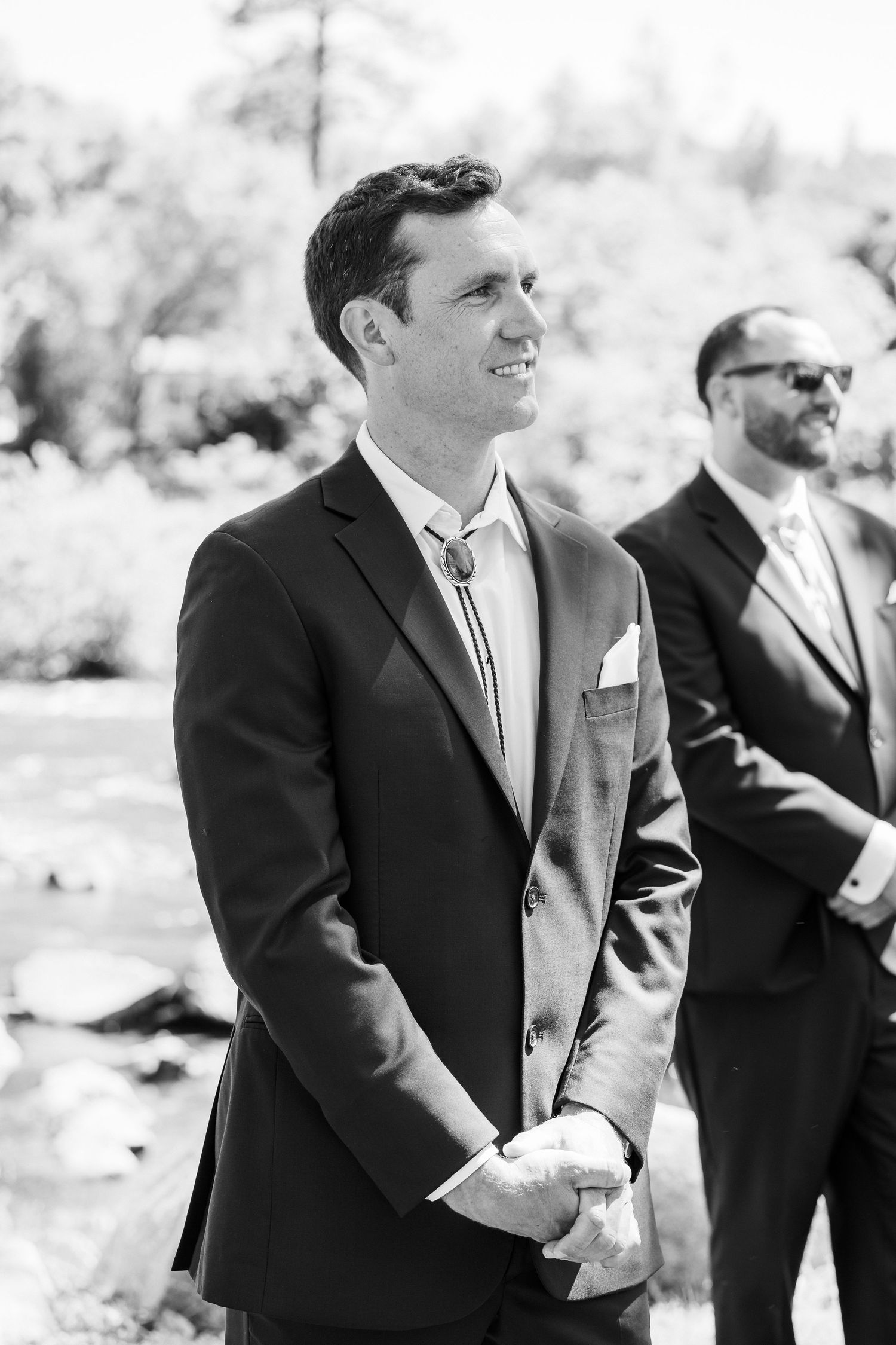 A group of groomsmen in black suits share a candid moment of laughter at an outdoor wedding ceremony.