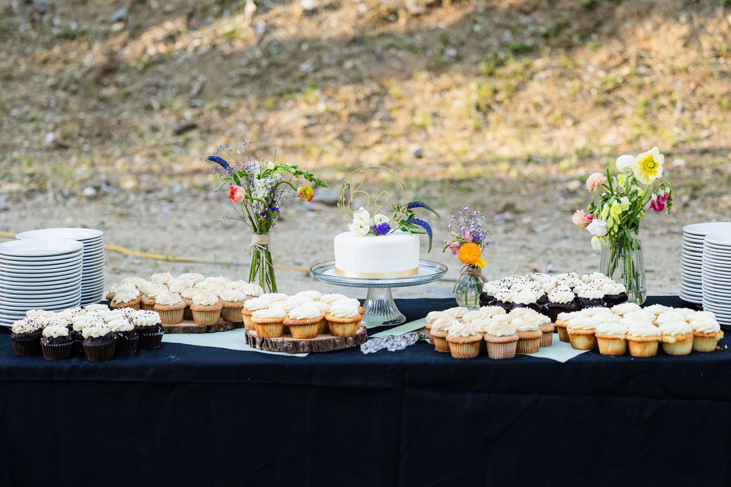 A panoramic view of dessert catering setup with cupcakes and flowers arranged on rustic wooden tables outdoors.