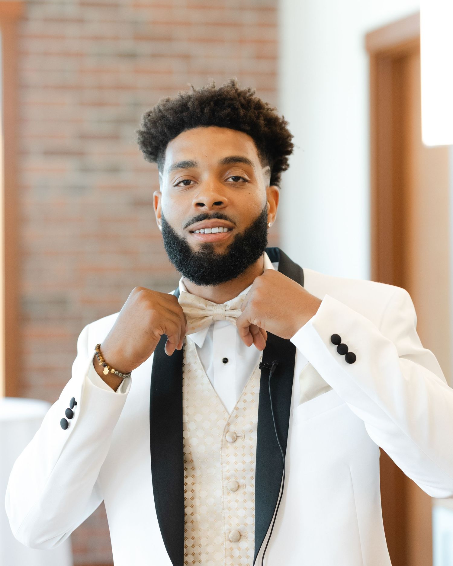 Groomsman in white dress shirt and gold vest smiles while adjusting his tie in a brick-walled room.