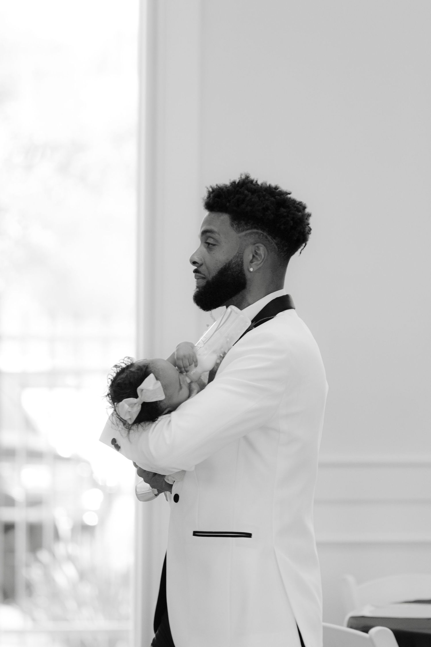 Groom in white tuxedo jacket and black bow tie stands in profile against bright white background.