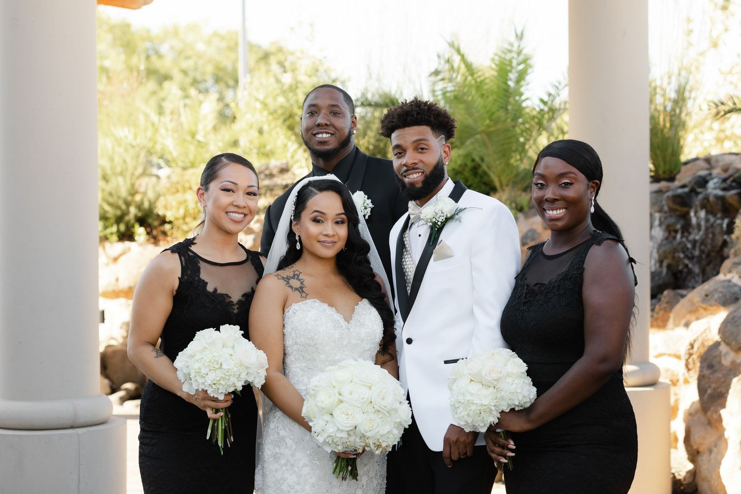 Wedding party members in formal attire pose together with white floral bouquets in an outdoor setting with columns.