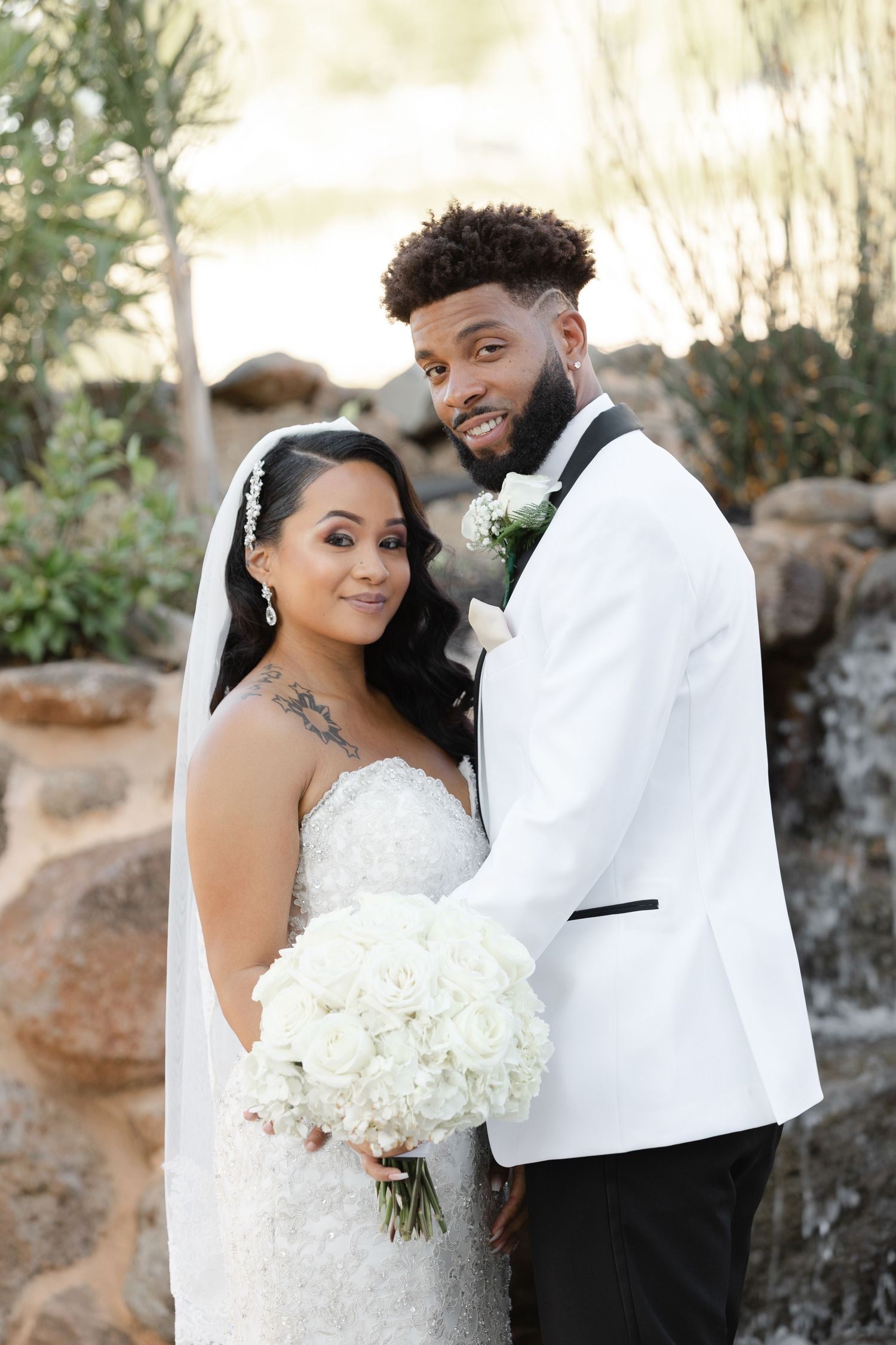 A romantic wedding photoshoot featuring a couple in formal attire with a white bouquet against a stone wall backdrop.