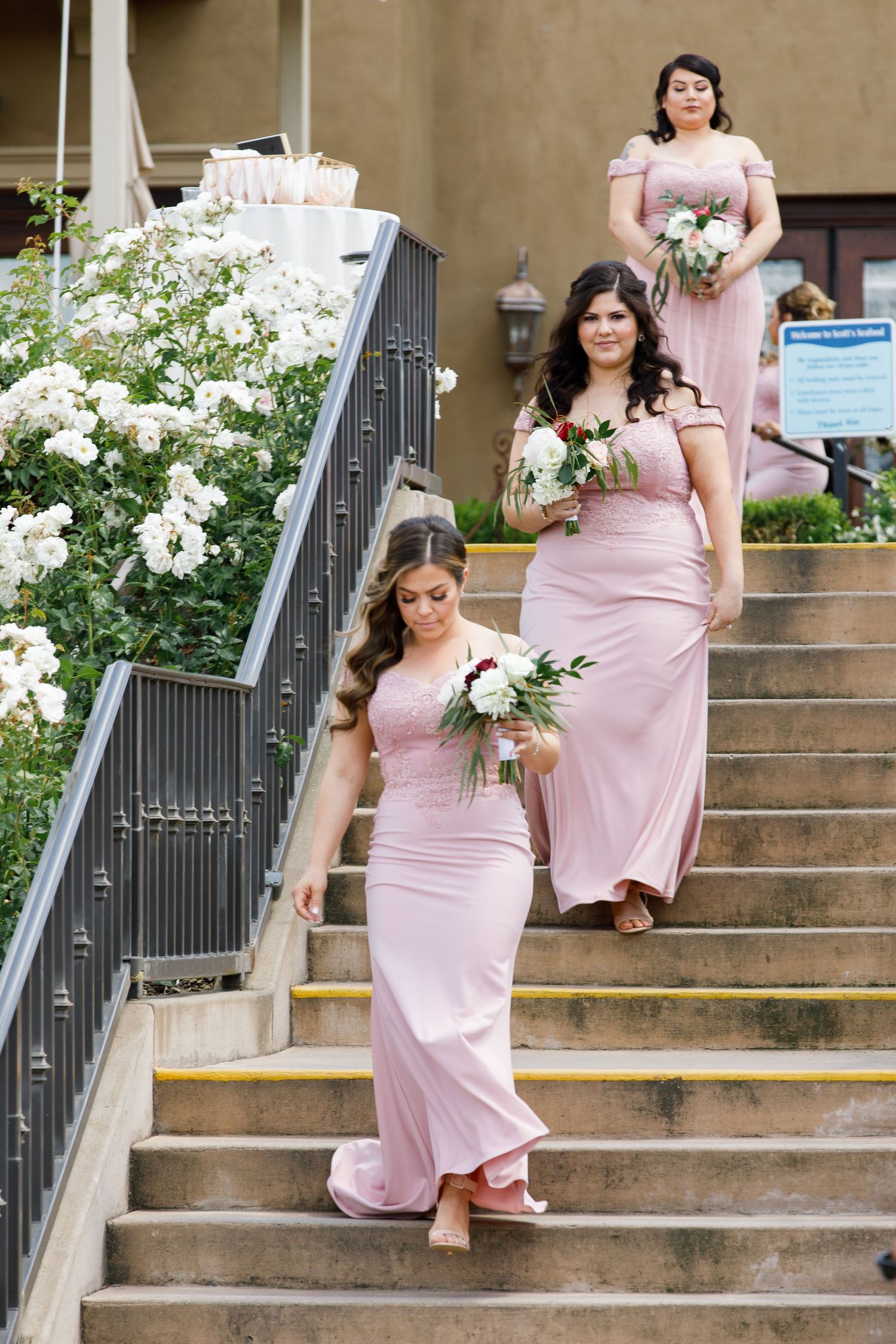 Bridesmaids in blush pink dresses sit gracefully on stone steps with white floral bouquets.