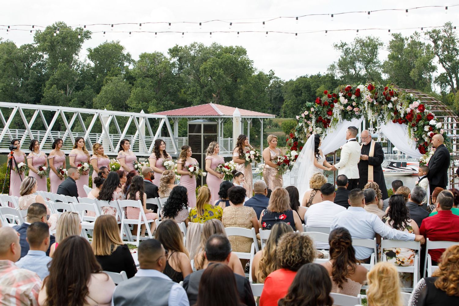 Guests stand during an outdoor wedding ceremony with white lattice arch and trees in the background.