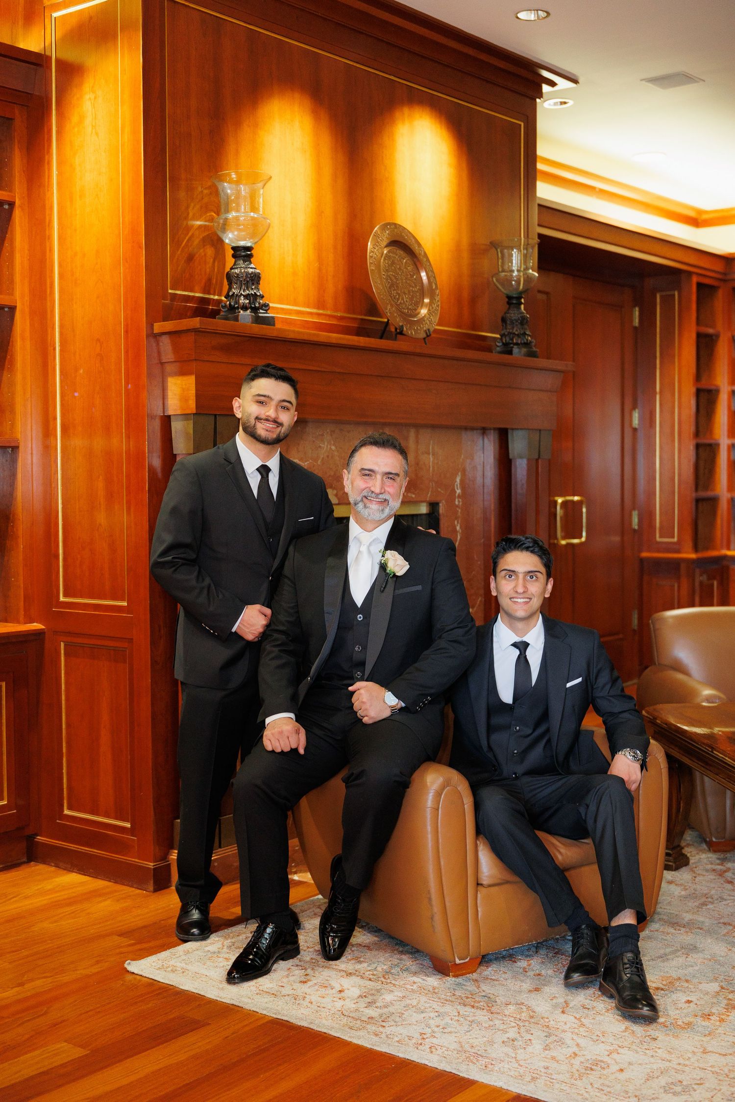 Three people in formal black suits and ties pose in an elegant wood-paneled room with leather furniture and decorative shelving.