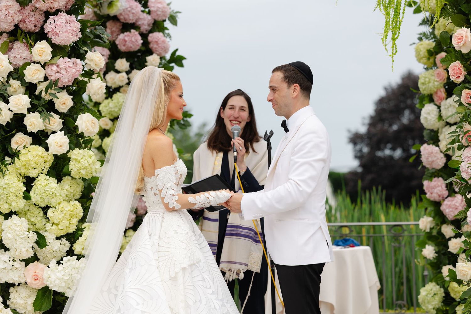 A romantic wedding ceremony moment captured outdoors with pink and white floral arrangements framing the scene.
