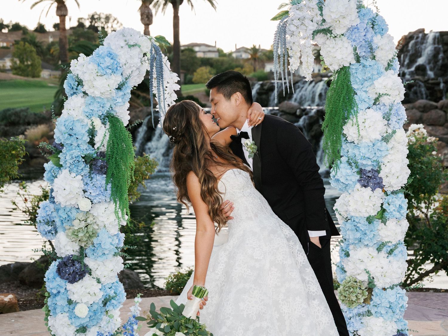 Romantic kiss under floral arch with blue and white flowers by waterside at outdoor wedding venue.