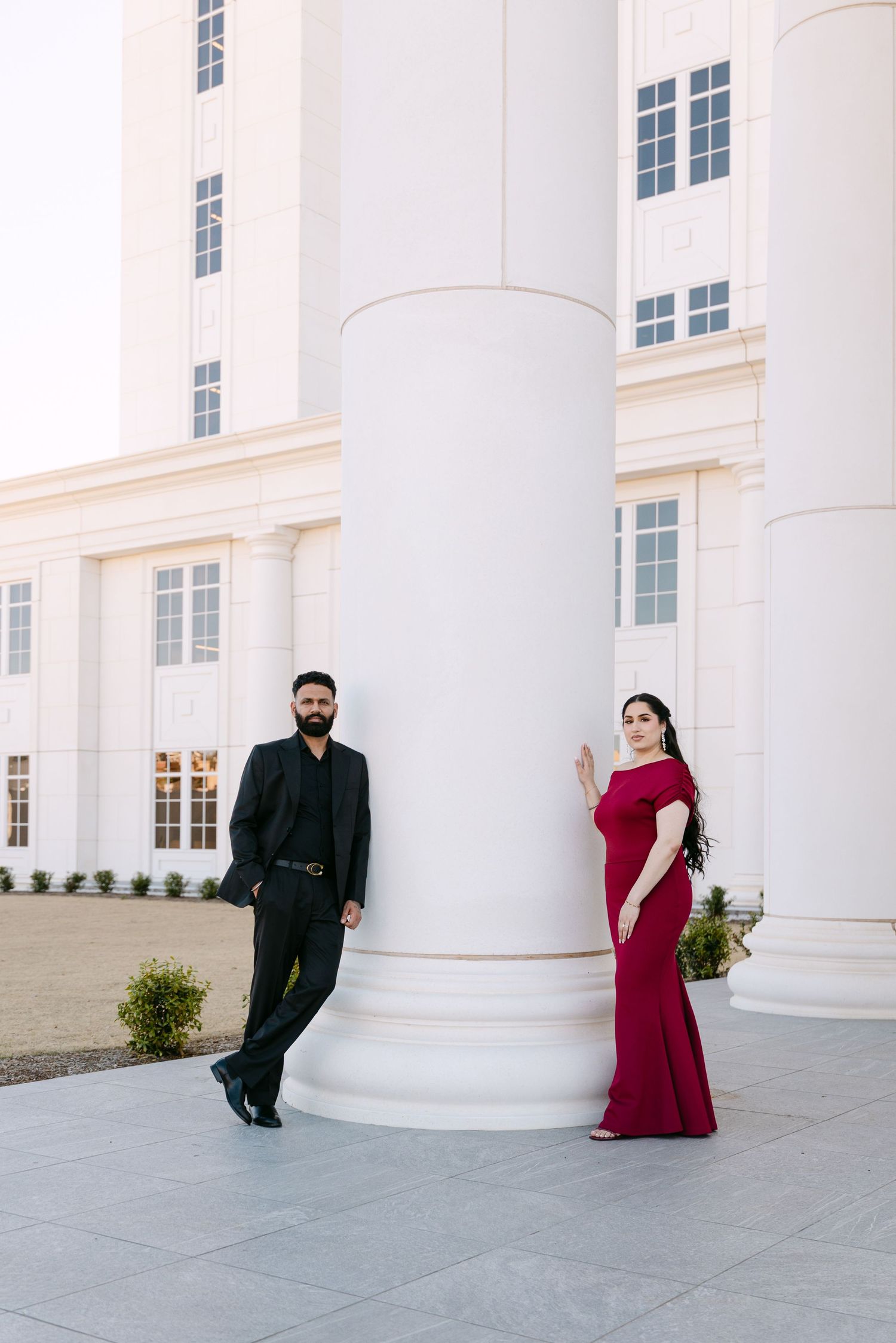 Elegant couple poses outside white columned building, with woman in red gown and man in black suit.