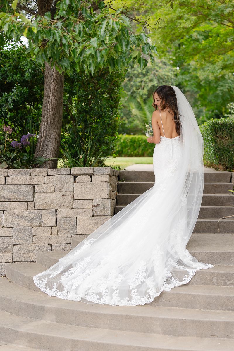 A flowing white wedding dress with lace trim and long train cascades elegantly down stone garden steps.
