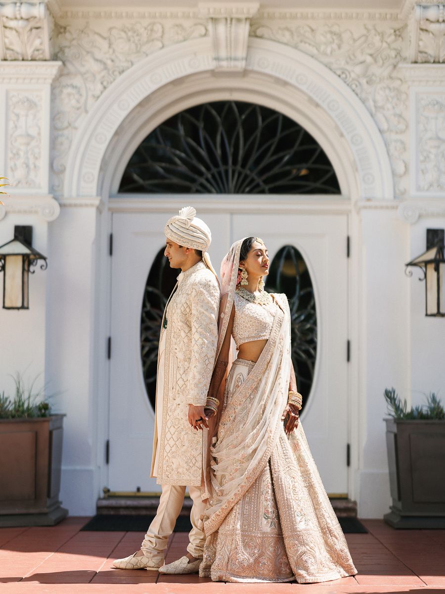 Couple in traditional South Asian wedding attire walking through elegant white archways.