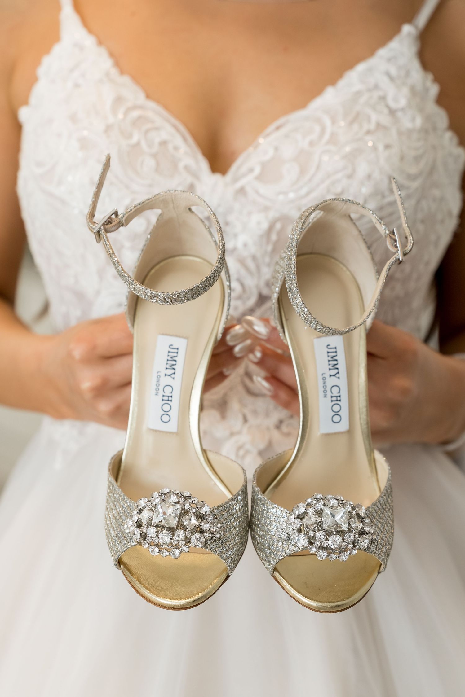 Elegant crystal-embellished silver bridal shoes with ankle straps displayed against white lace fabric.