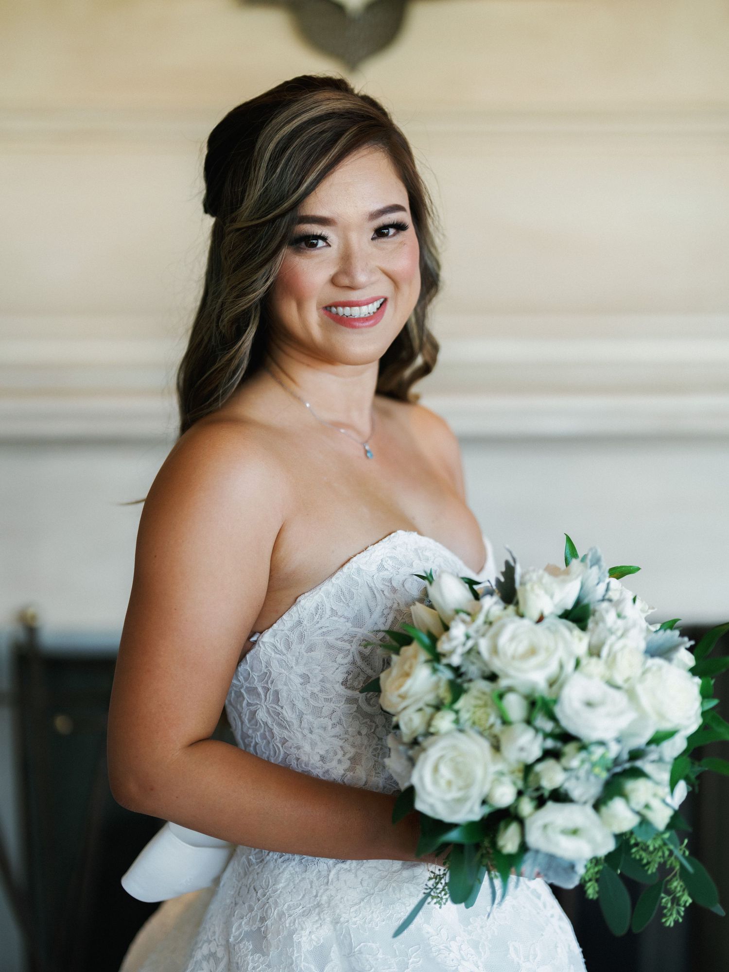 A bride in a strapless lace wedding gown holds a beautiful white rose bouquet with greenery.