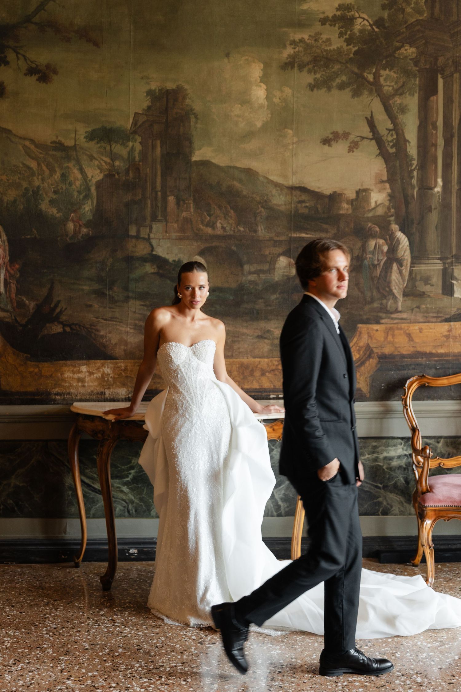 Elegant couple poses in formal wedding attire against a vintage mural backdrop in an ornate interior setting.