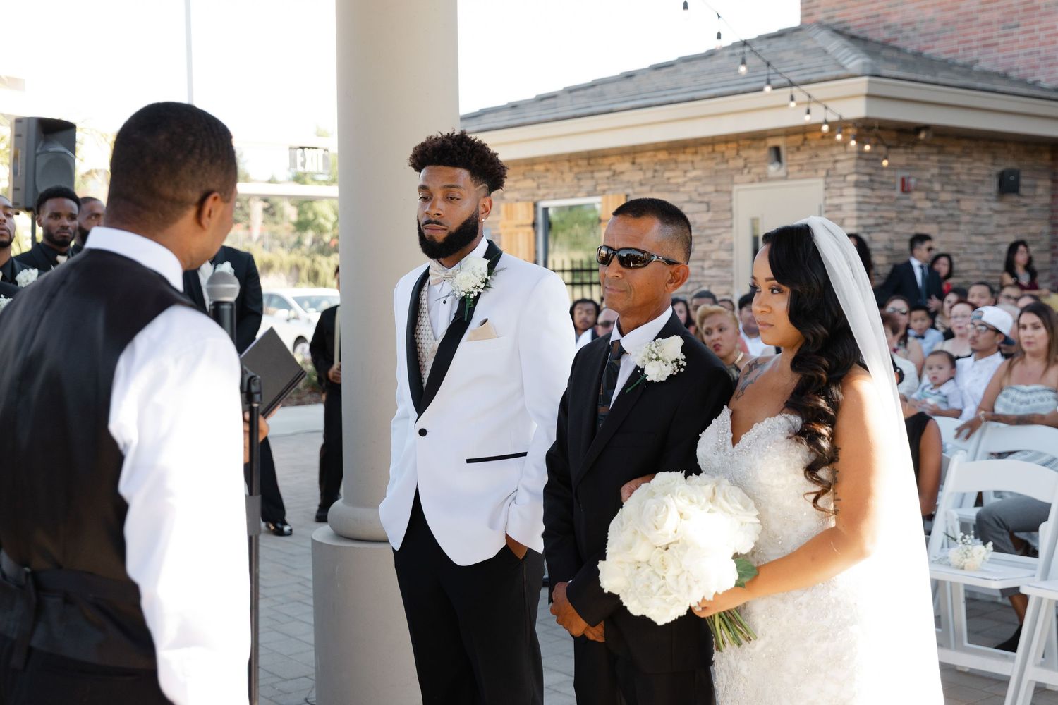 Wedding party members celebrate during outdoor reception with string lights and brick building in background.
