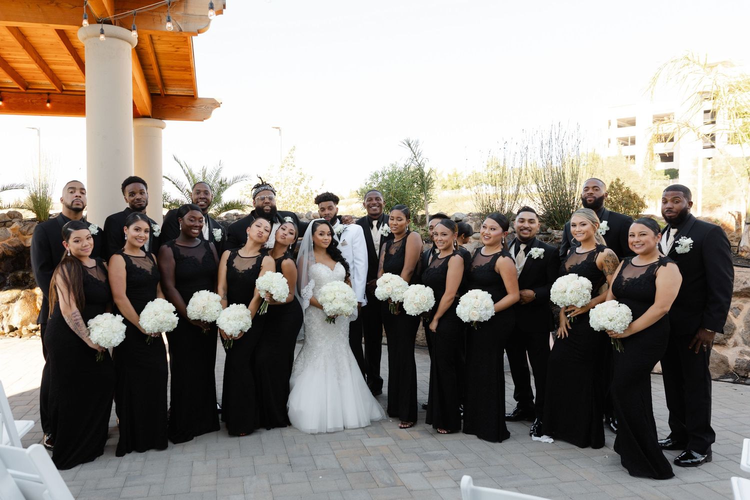 Wedding party wearing black and white formal attire poses together outdoors for group photos at sunset.