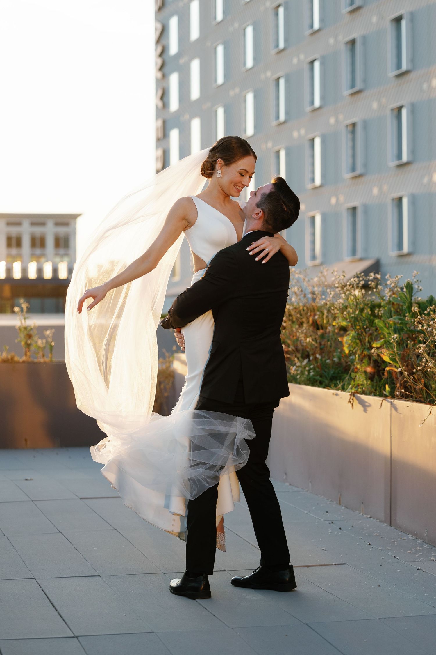 A couple in wedding attire dances and dips romantically on a rooftop terrace at sunset.