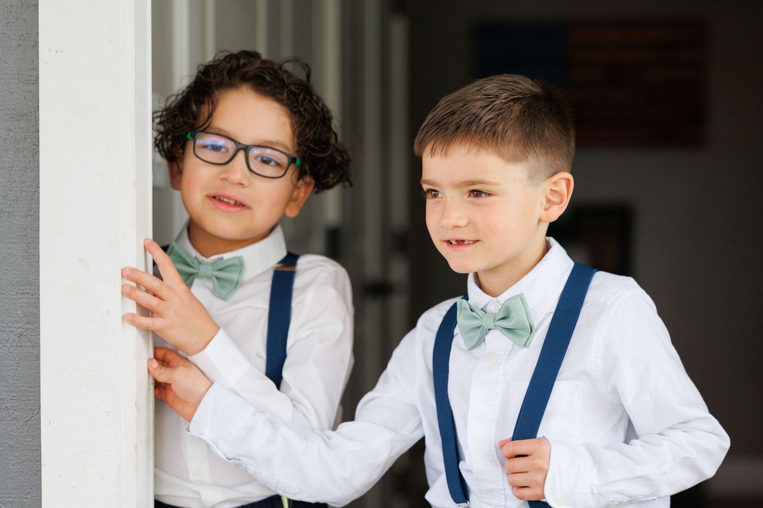 Two young children in white shirts and blue suspenders smile and laugh while posing together.
