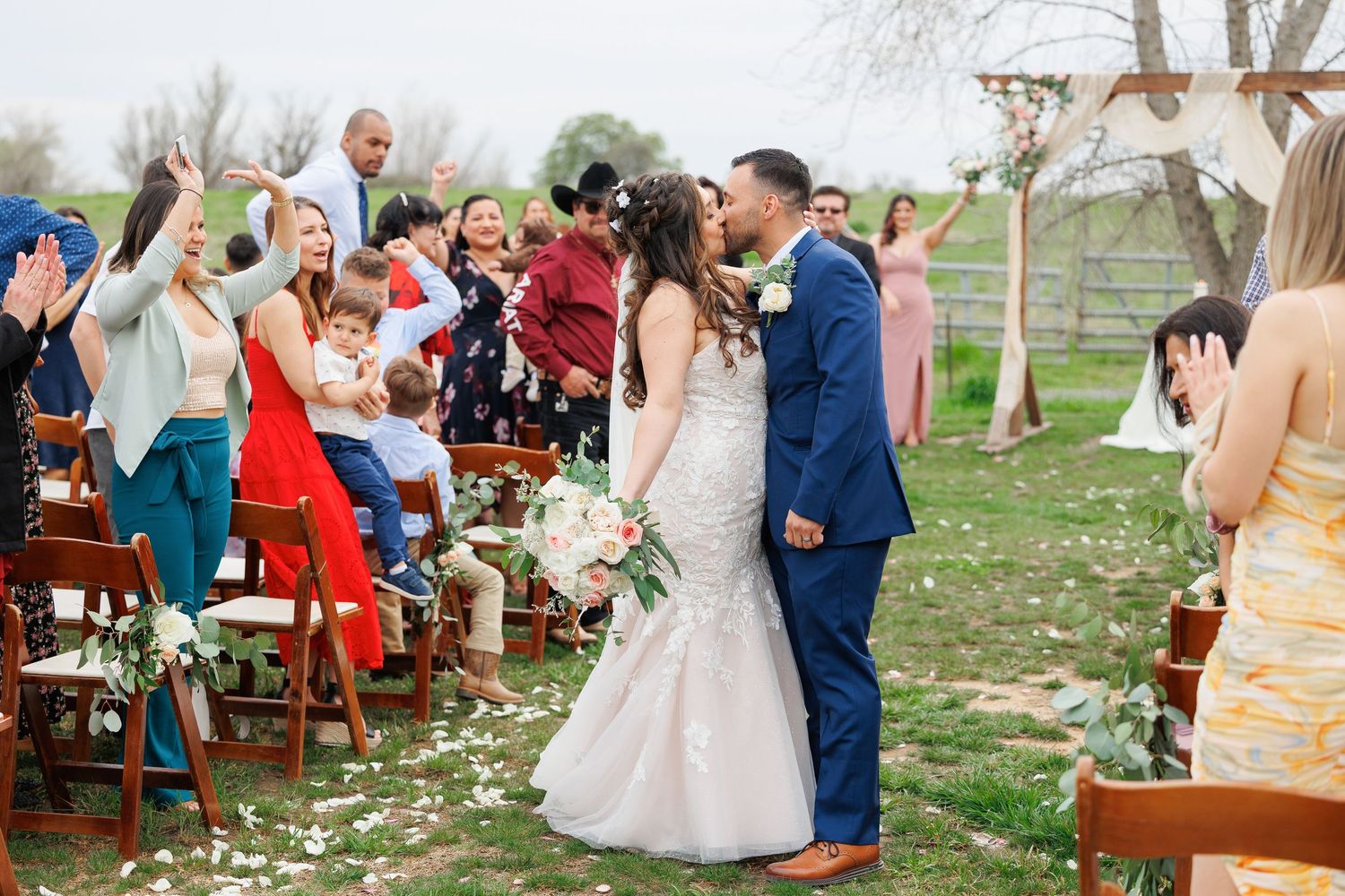 Wedding guests celebrate outdoors at a rustic reception with wooden tables and chairs under a sunny sky.
