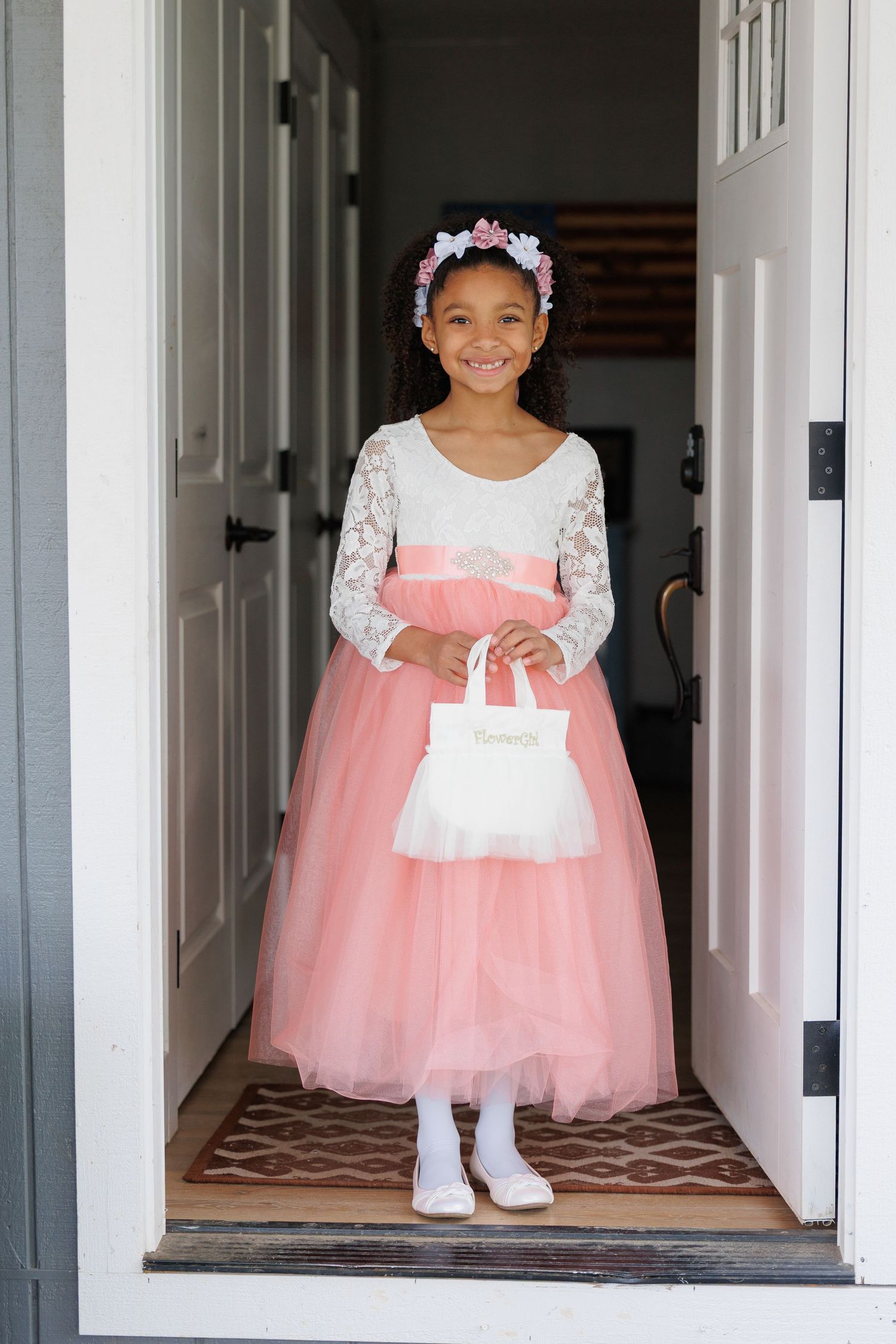 A young girl in a pink tulle dress and white lace stands in a doorway wearing a flower headpiece.