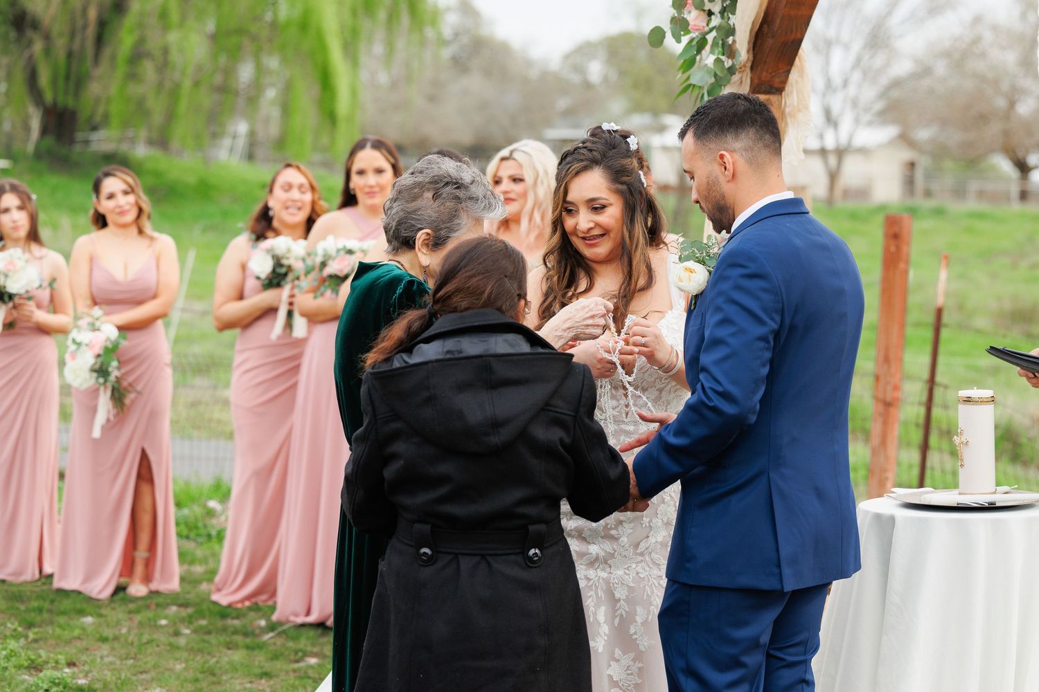 A couple exchanges vows during an outdoor wedding ceremony with bridesmaids in pink dresses looking on.