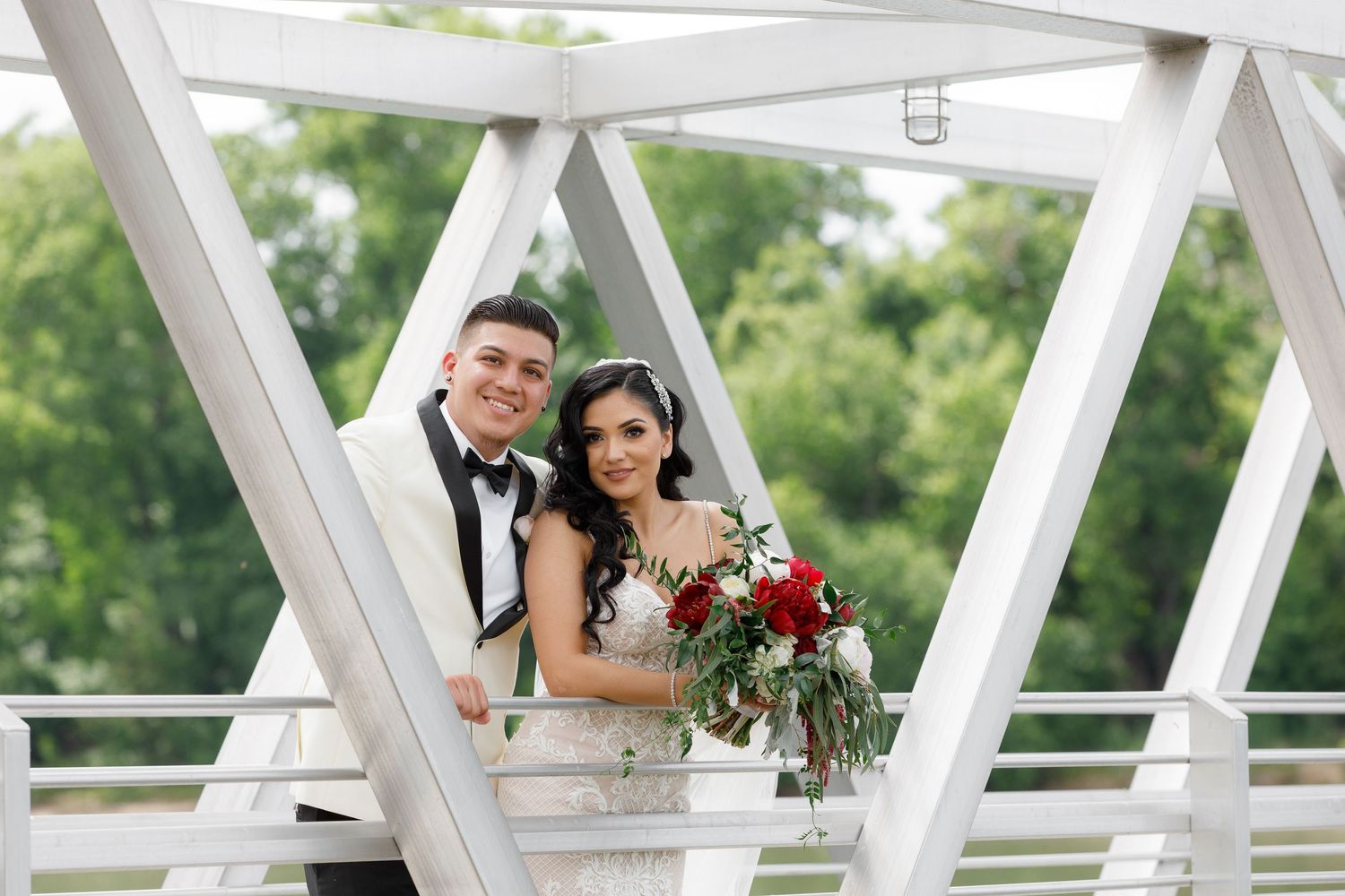 Newlyweds pose on a white wooden bridge surrounded by lush greenery during an outdoor wedding photo session.