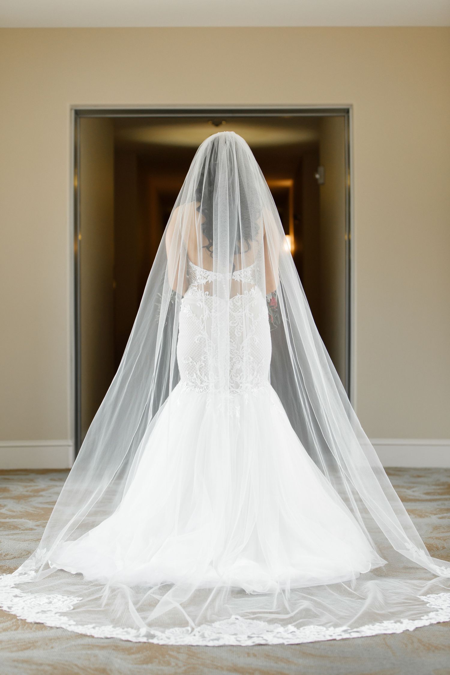 A bride in a flowing white wedding dress and cathedral veil sits elegantly on the floor in a dramatic silhouette pose.