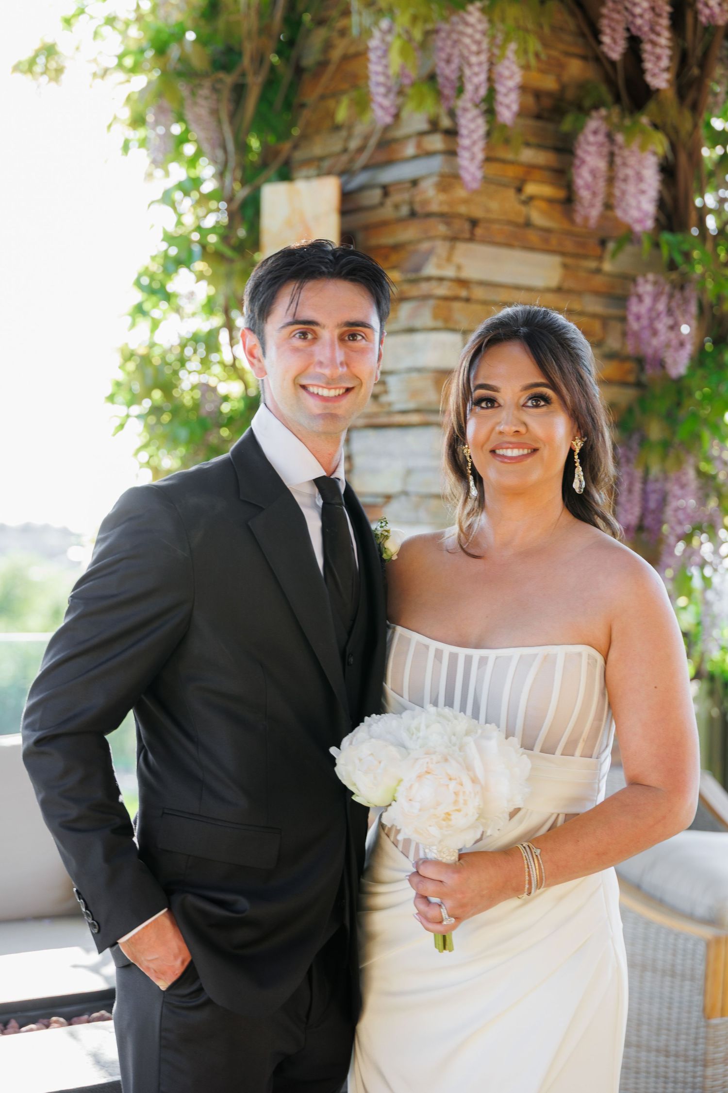 A happy couple shares joyful moments in formal wedding attire under a wisteria-covered pergola.