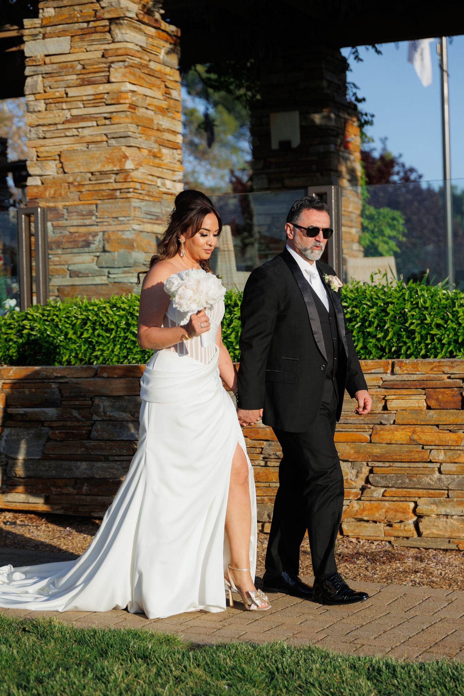 A couple in formal wedding attire walks along a stone-lined pathway outside a building.