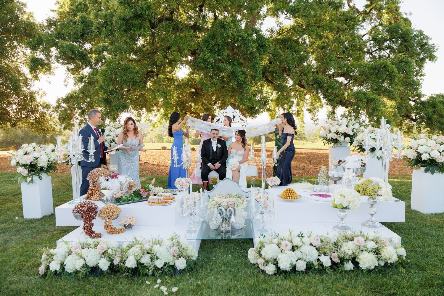Wedding reception guests gather around elegant white table settings on a lush green lawn at sunset.