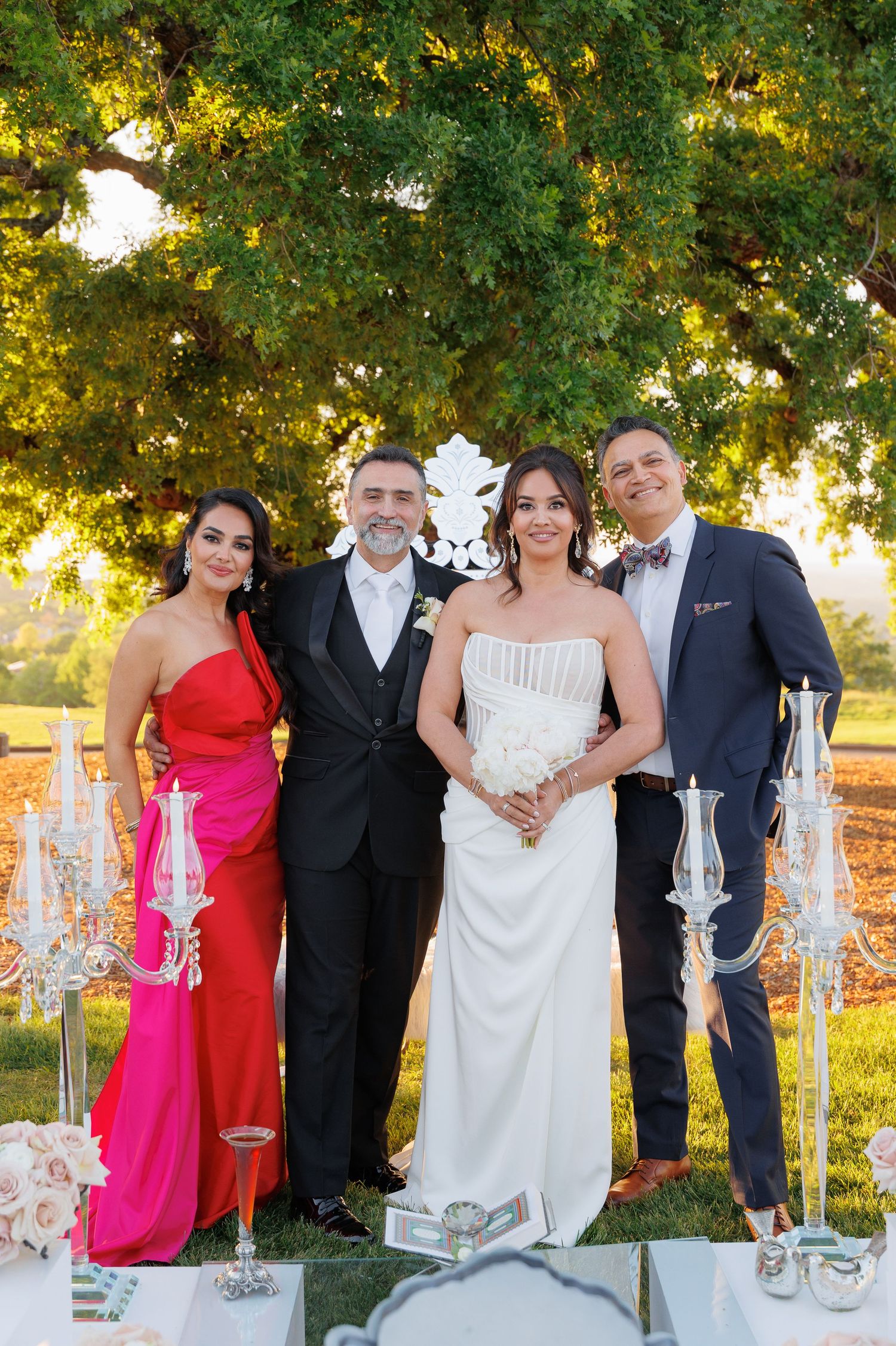 Elegant wedding party poses outdoors with champagne glasses and decorative candles at sunset under a lush green tree.
