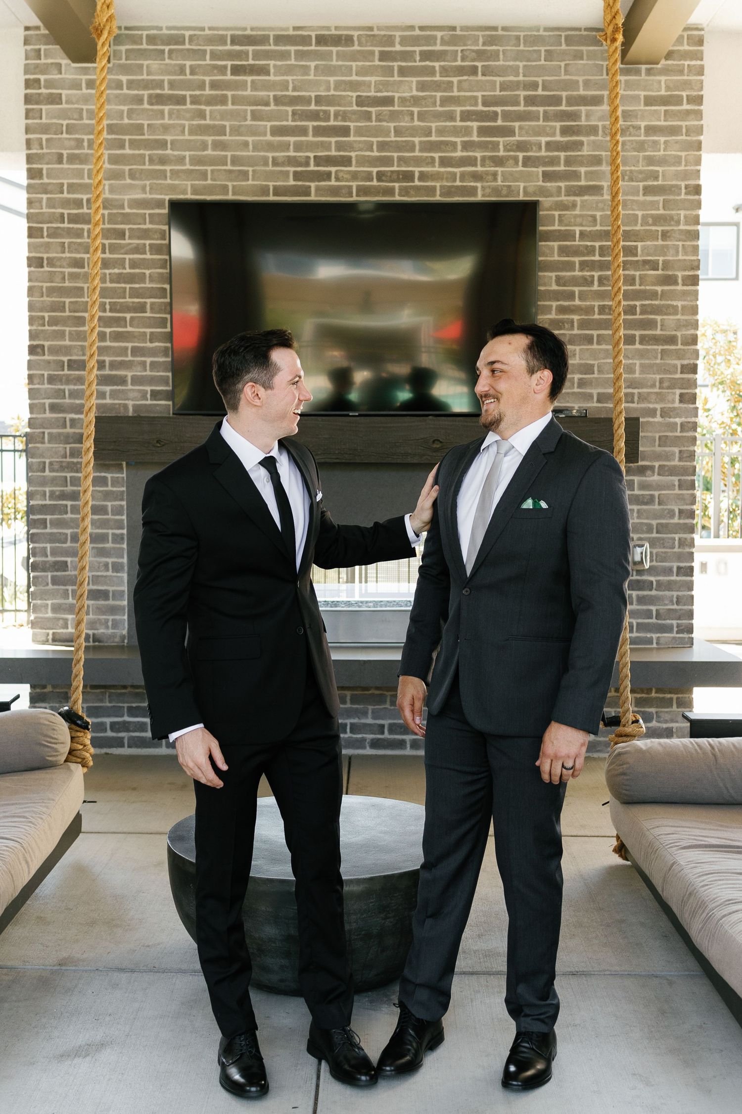 Two people in black suits share a candid moment while standing in front of a brick wall.