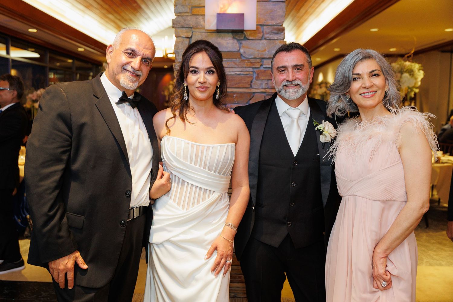 Wedding party members pose together in formal attire at an indoor venue with warm lighting and stone walls.