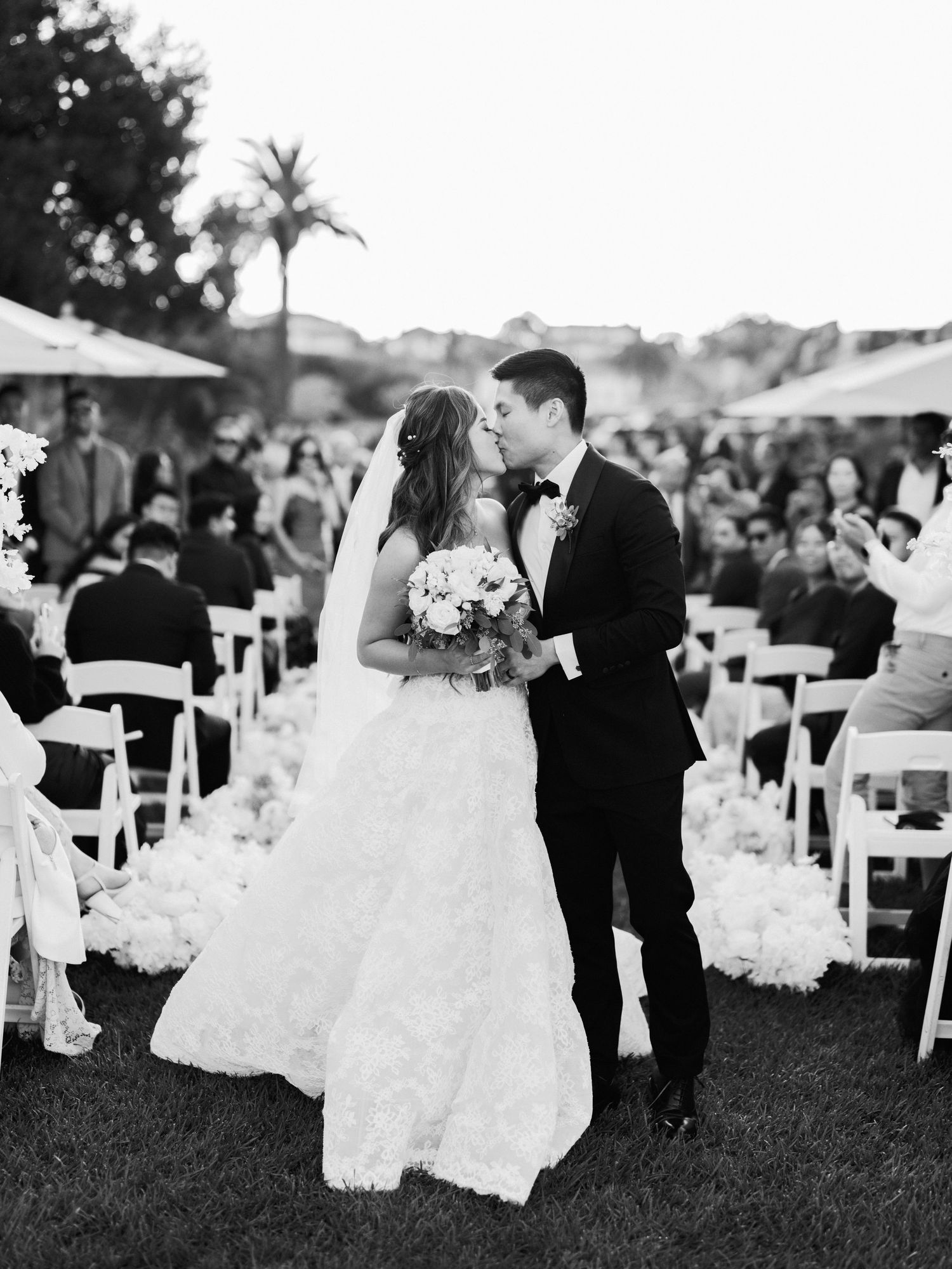A black and white outdoor wedding ceremony photo with white chairs and palm trees.
