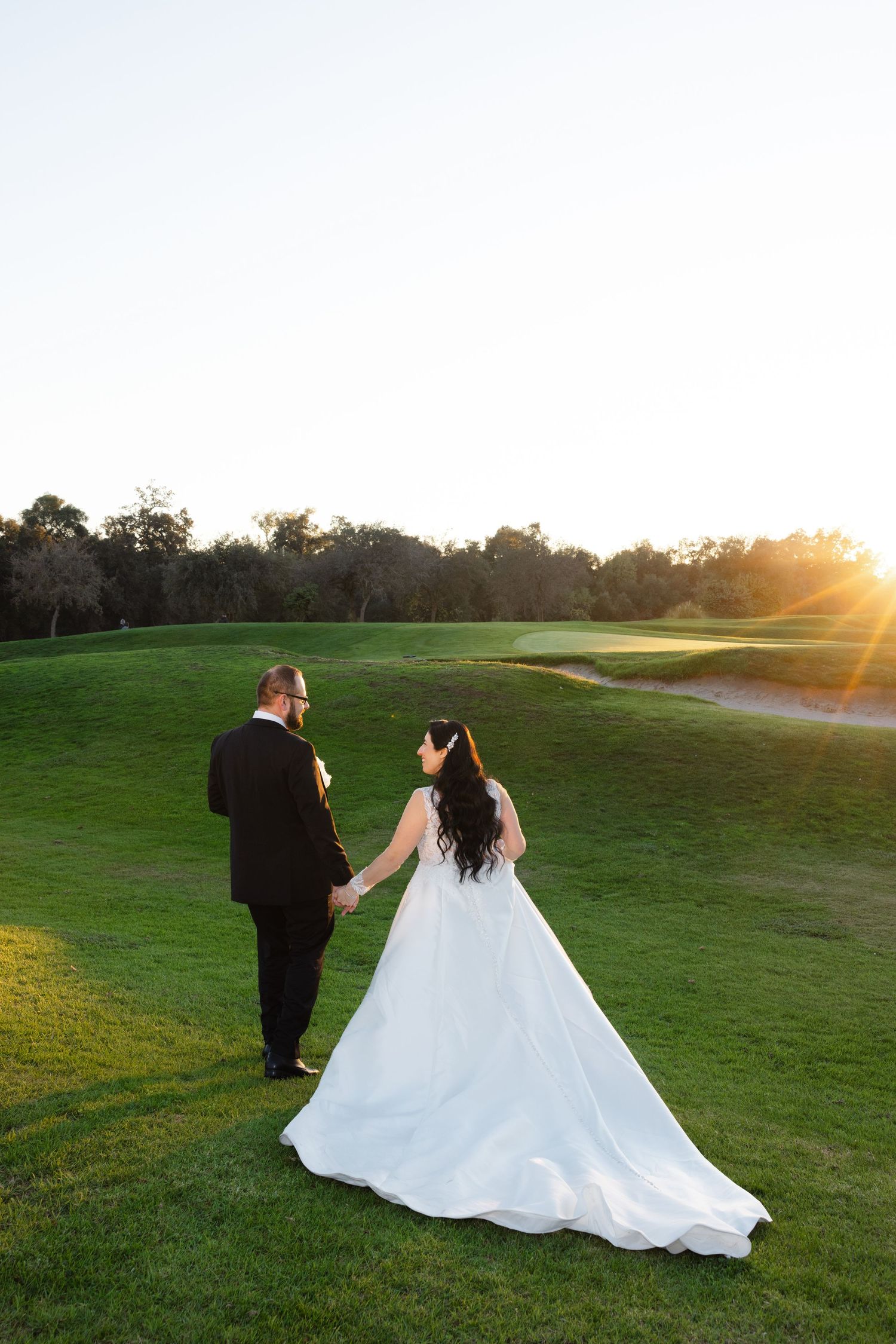 A couple in wedding attire walks hand in hand on a golf course at sunset with golden sunlight streaming through trees.