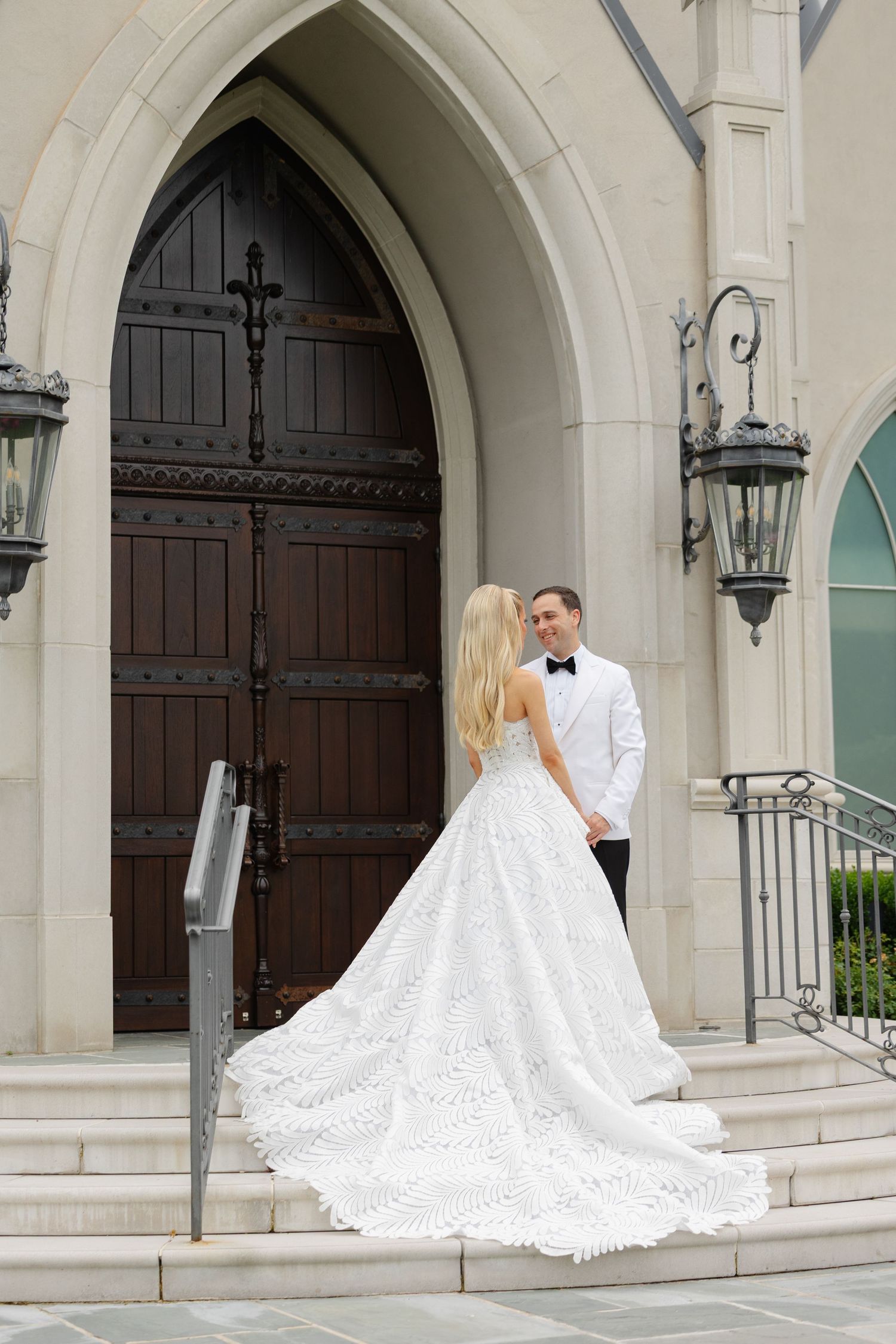 Bride in white lace gown with long train stands with groom in white tuxedo on church steps before ornate wooden doors.