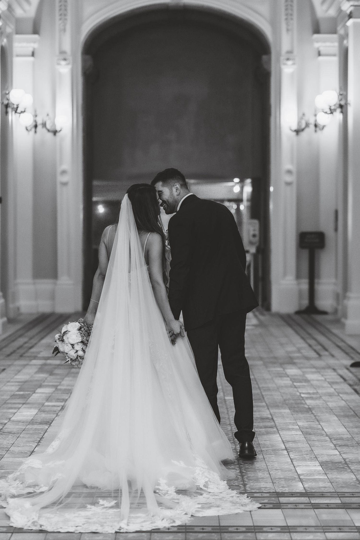 Black and white photo of bride and groom kissing in elegant hall, bride wearing long veil and gown holding bouquet.
