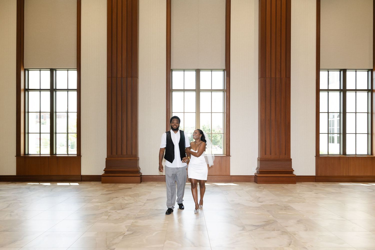 Couple posing in elegant ballroom with tall windows and columns on marble floor.