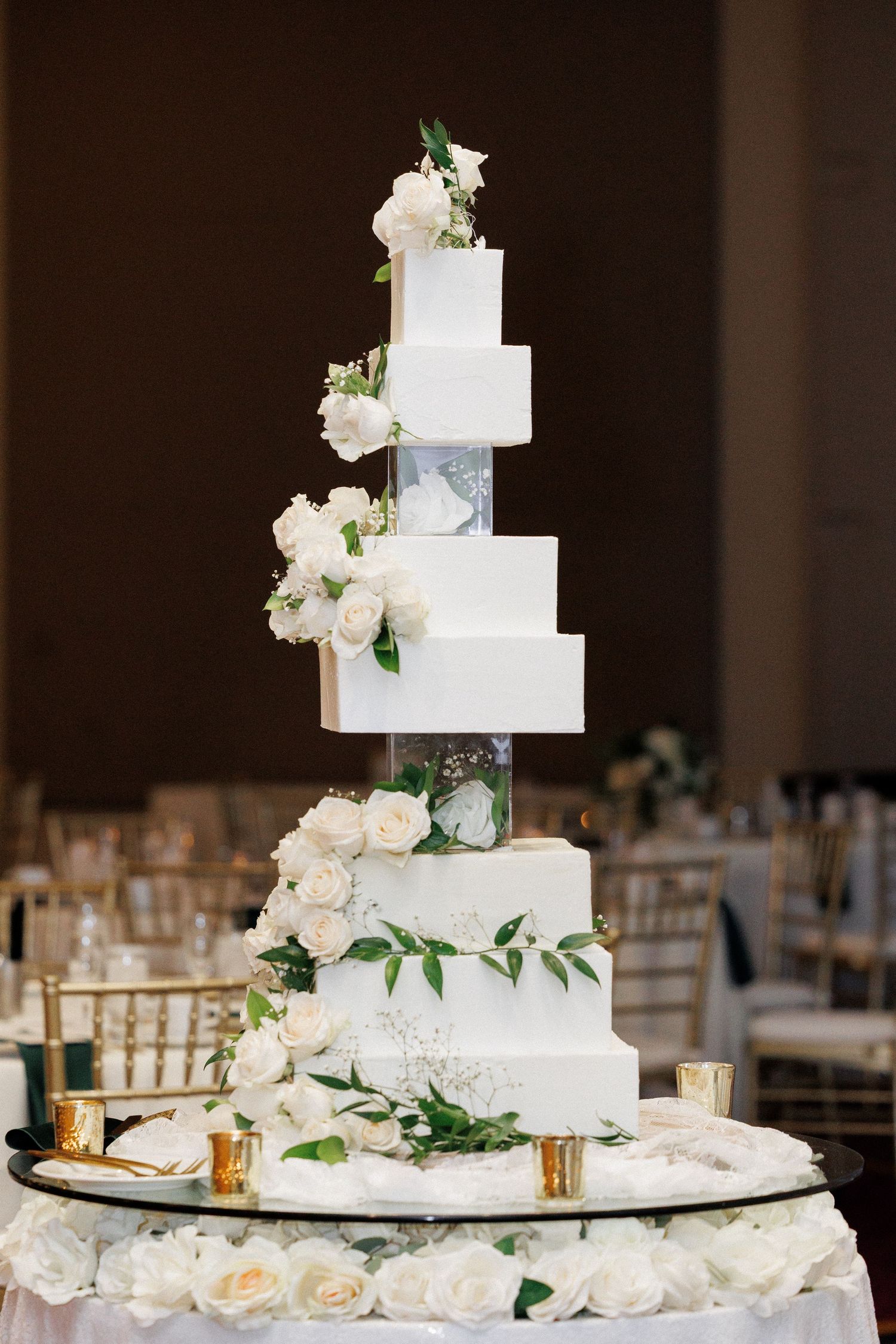 Elegant six-tier white wedding cake decorated with fresh white roses and greenery on marble display stand.
