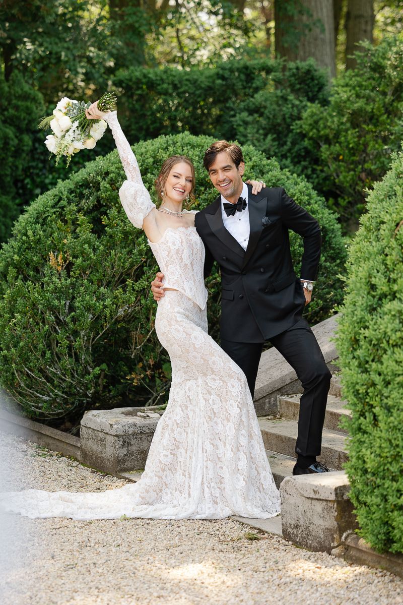 A couple in wedding attire dances playfully on a garden path surrounded by lush greenery.