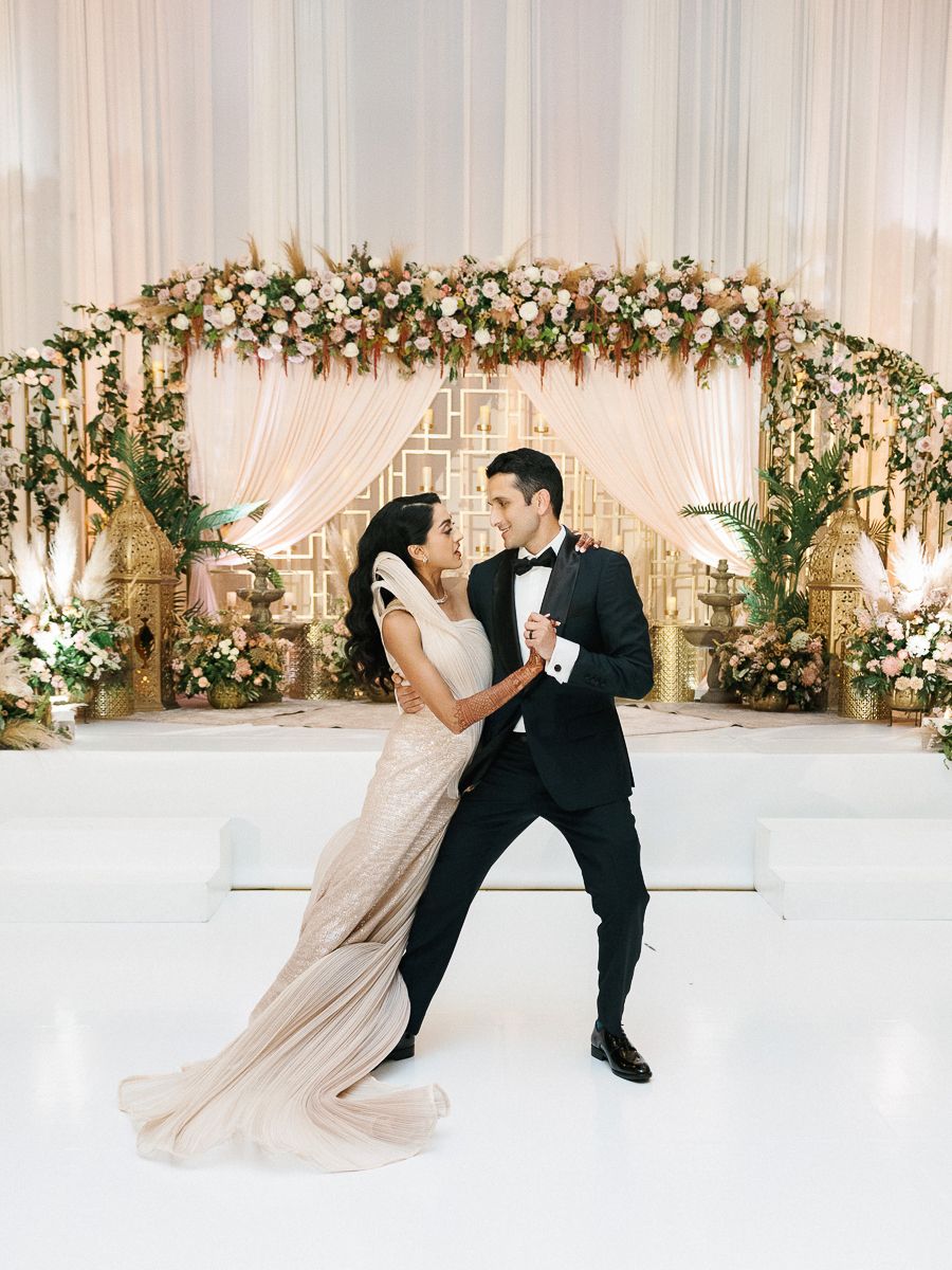 Couple dancing together on white floor with elegant draped backdrop and floral decorations.