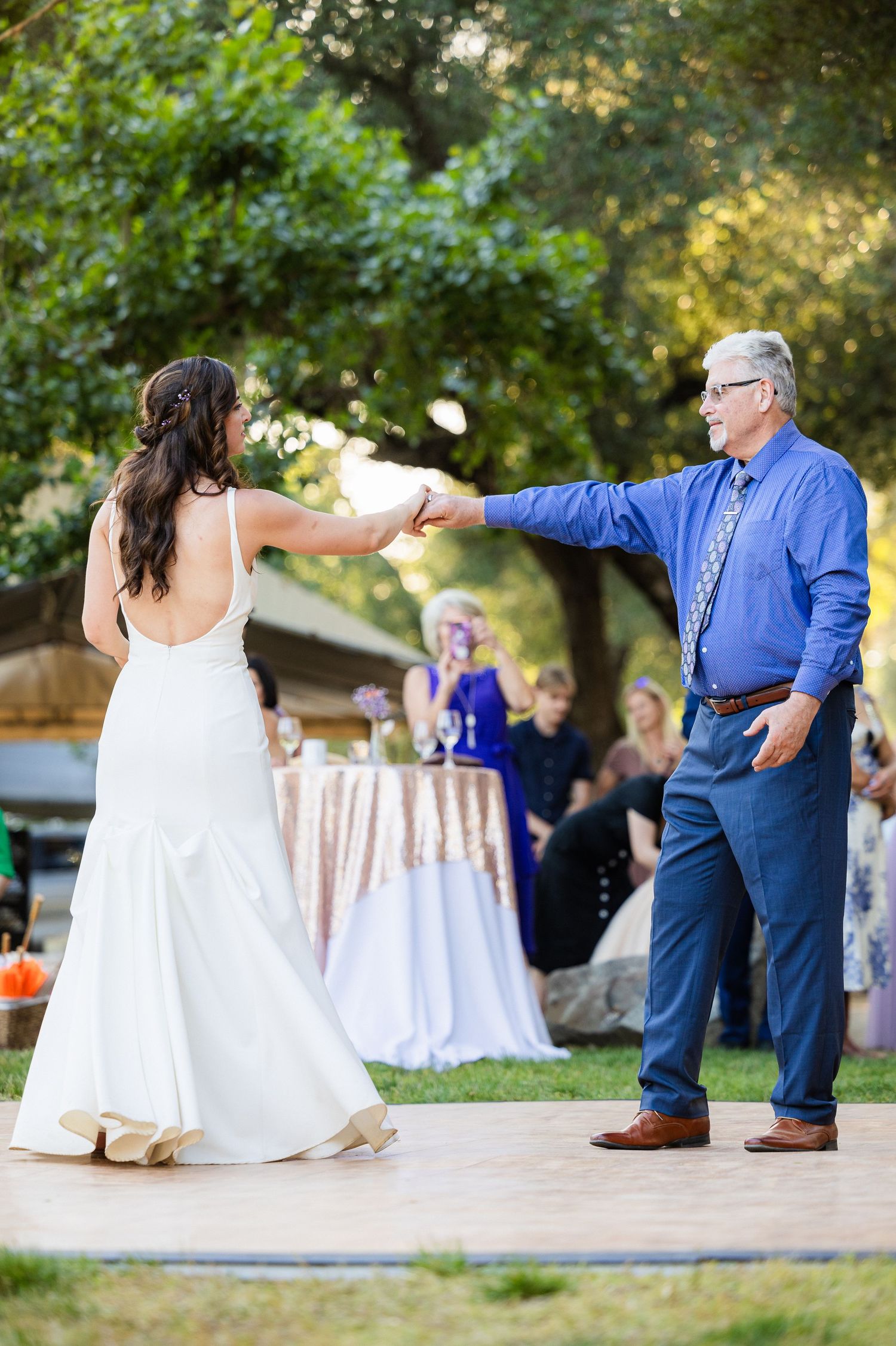 A couple dances together during an outdoor wedding reception on a wooden dance floor with decorative tables in background.