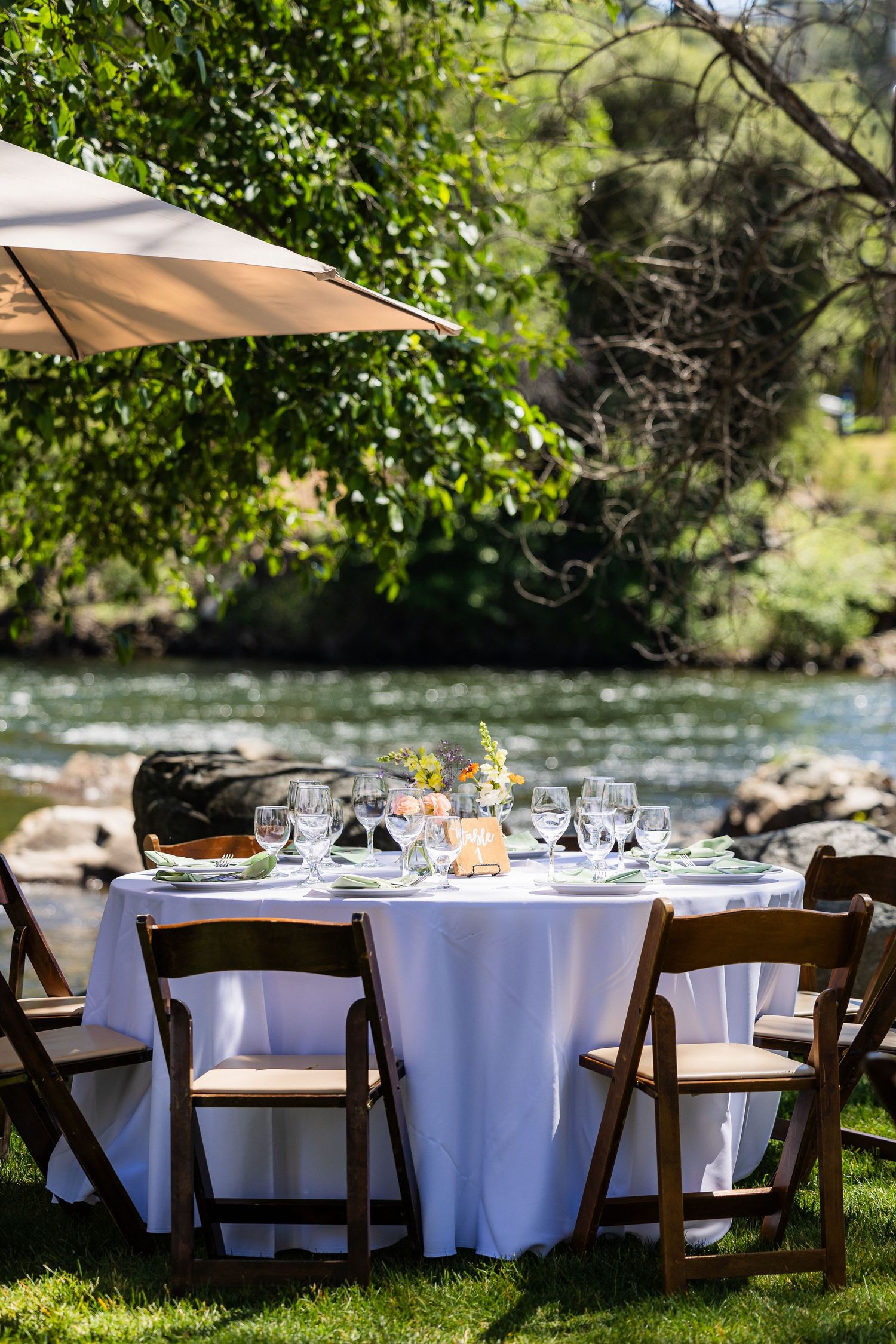 Elegant outdoor wedding reception setup with round tables covered in white linens and wooden chairs by a river.