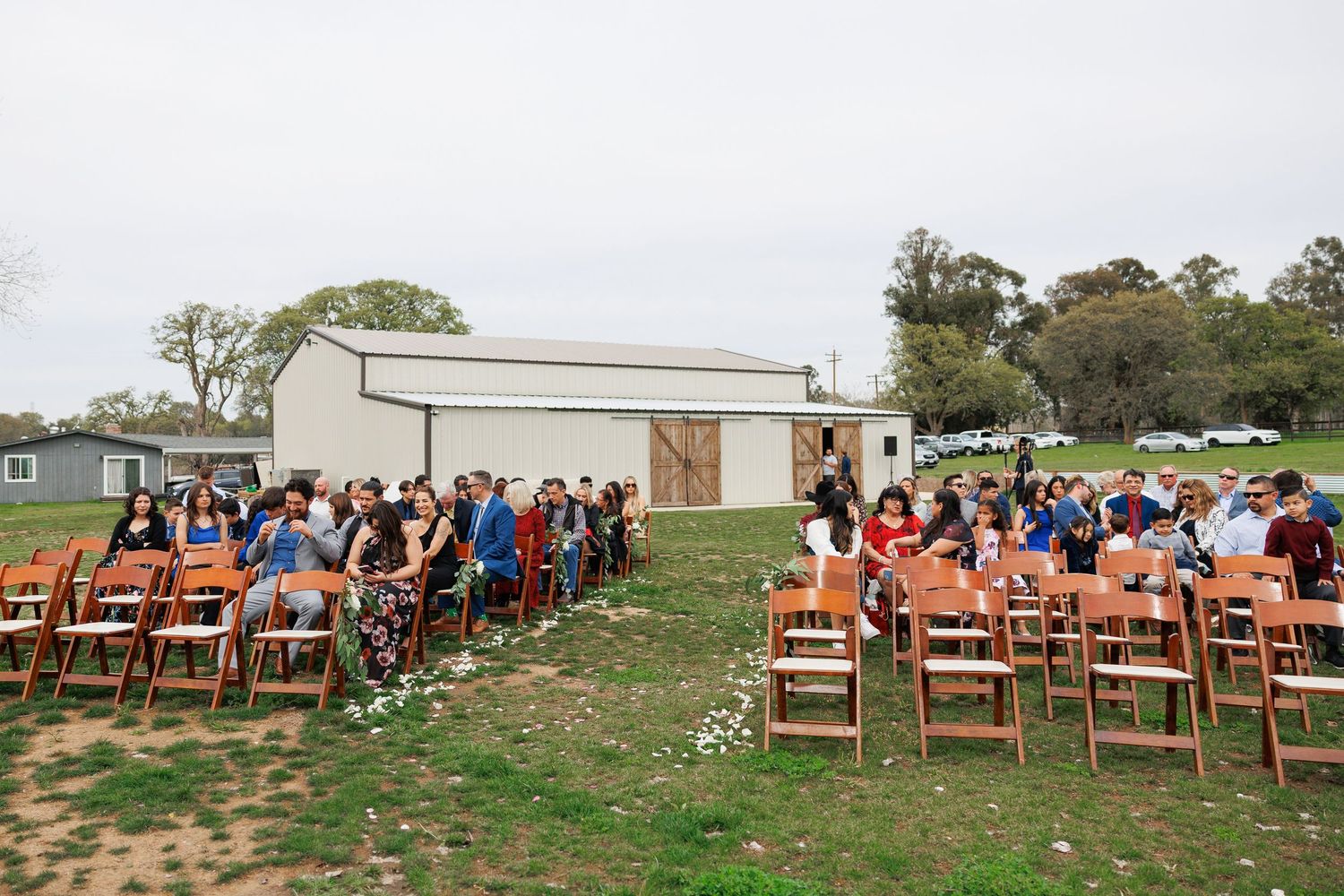 Wedding guests seated in wooden chairs on a grassy lawn outside a white barn venue for an outdoor ceremony.