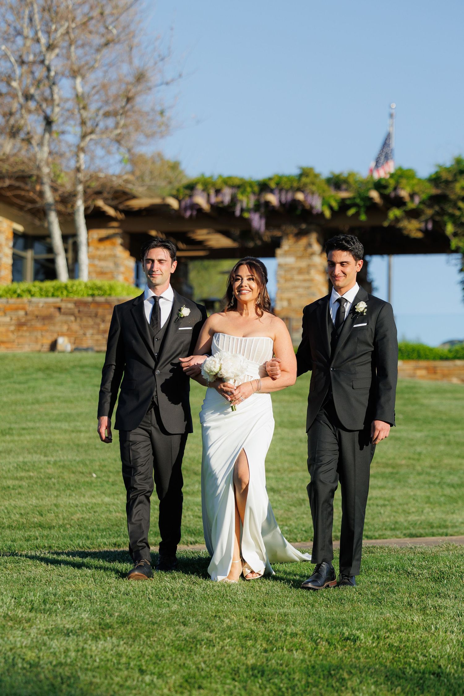 Wedding party walks across green lawn during outdoor ceremony in late afternoon sunlight.