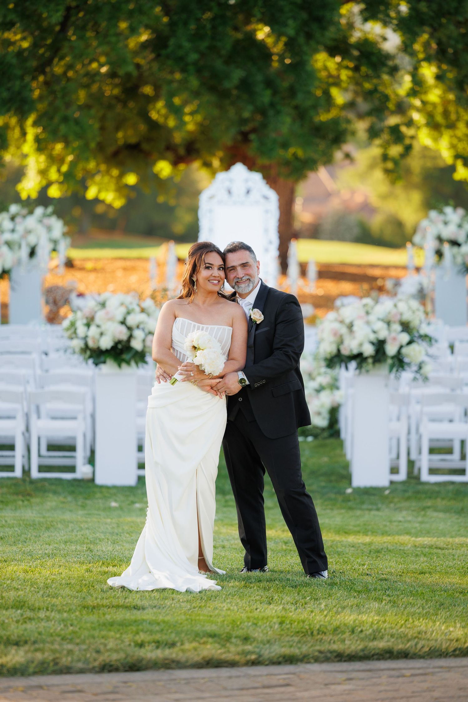 Outdoor wedding ceremony setup with white chairs and floral arrangements under golden sunlight in a garden setting.